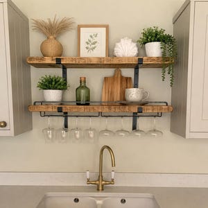 May include: A kitchen shelf unit with two reclaimed wooden shelves and black metal supports. The top shelf features a framed print, a vase, and a shell. The lower shelf holds wine glasses, a wooden chopping board, and a green bottle.