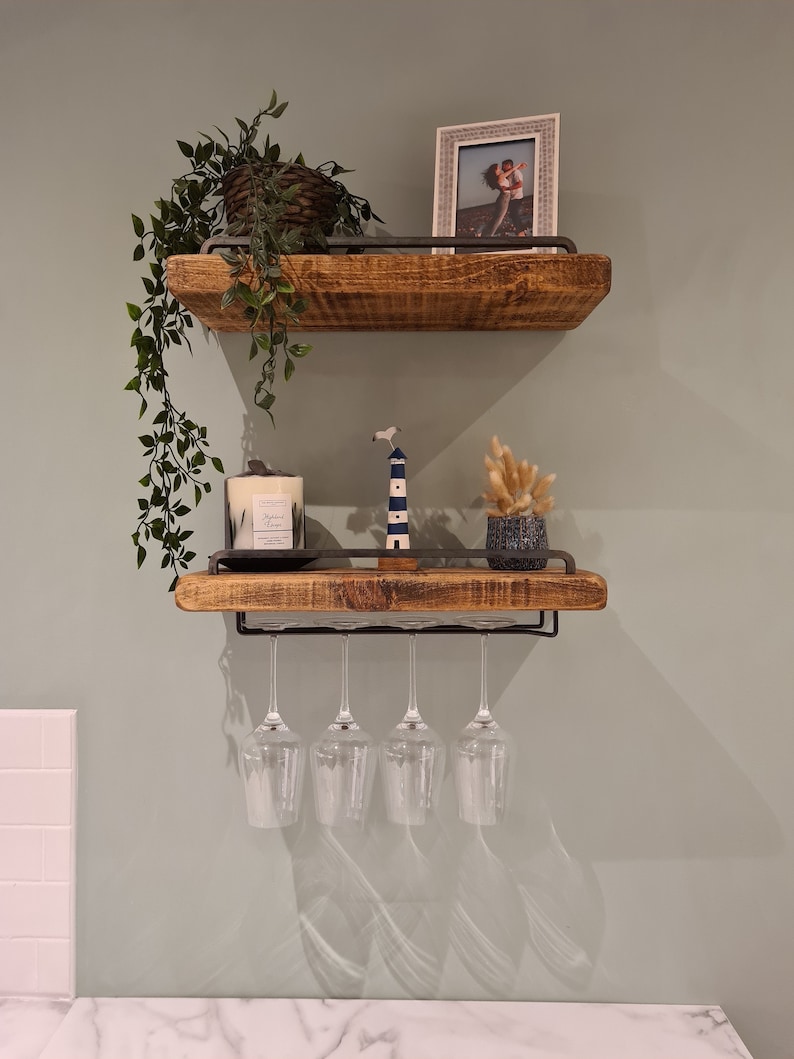 K&ouml;nnte beinhalten: Zwei Holzregale mit einem Metallrahmen, montiert an einer hellgr&uuml;nen Wand. Das obere Regal hat eine Topfpflanze und ein gerahmtes Foto. Das untere Regal hat eine Kerze, eine kleine Leuchtturmfigur, eine kleine Topfpflanze und einen Weinglashalter mit vier leeren Gl&auml;sern.