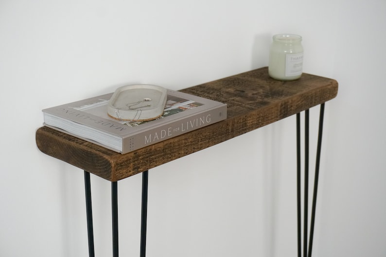 May include: A rustic wooden console table with a distressed brown finish. The table has a narrow top and three black hairpin legs. A book titled "Made for Living" and a decorative tray are on the table.