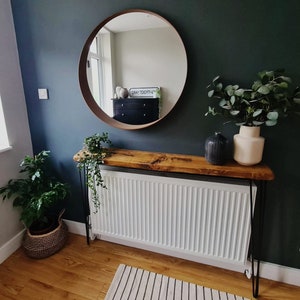 May include: A wooden console table with black hairpin legs, a white radiator, and a round mirror with a brown frame. The table is decorated with a potted plant, a vase with green leaves, and a black decorative object. The wall is painted a dark green color.