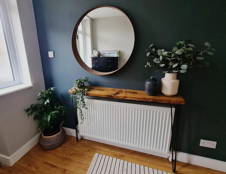 May include: A wooden console table with black hairpin legs, a white radiator, and a round mirror with a brown frame. The table is decorated with a potted plant, a vase with green leaves, and a black decorative object. The wall is painted a dark green color.