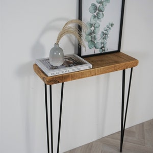 May include: A rustic wooden console table with a natural wood finish and black hairpin legs. The table is decorated with a book, a vase, and a framed print of eucalyptus leaves.