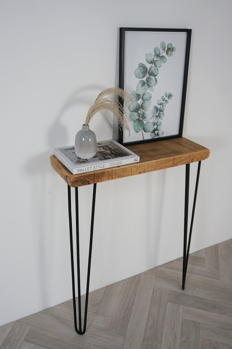 May include: A rustic wooden console table with a natural wood finish and black hairpin legs. The table is decorated with a book, a vase, and a framed print of eucalyptus leaves.