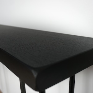 K&ouml;nnte beinhalten: Schwarzer Konsolentisch mit einer dicken, rechteckigen Tischplatte und einem schlichten, modernen Design. Der Tisch hat ein schwarzes Metallgestell mit d&uuml;nnen Beinen.