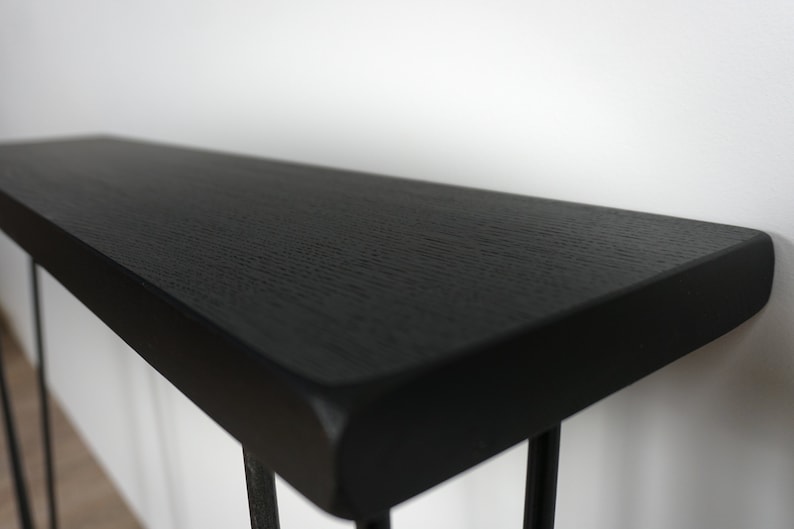 K&ouml;nnte beinhalten: Schwarzer Konsolentisch mit einer dicken, rechteckigen Tischplatte und einem schlichten, modernen Design. Der Tisch hat ein schwarzes Metallgestell mit d&uuml;nnen Beinen.