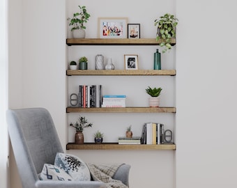 May include: A built-in wooden shelving unit with four shelves, displaying potted plants, books, decorative vases, and framed photos. A gray armchair with pillows and a blanket is in the foreground, creating a cozy interior design.