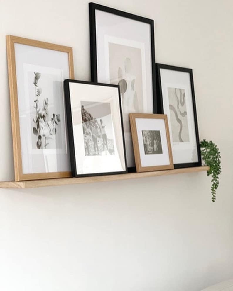 May include: A wooden shelf with five framed pictures, including black and white photos and abstract art. The shelf is mounted on a white wall with a small green plant hanging on the right side.