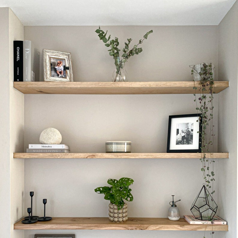 May include: A wooden shelf unit with three shelves, featuring various decorative items, including a framed photo, a vase with dried greenery, a candle, a book, and a small potted plant. The shelves are against a light beige wall.