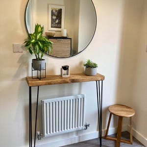May include: A wooden console table with black hairpin legs, a round mirror with a black frame, and a white radiator. There are two potted plants and a small wooden stool on the table.