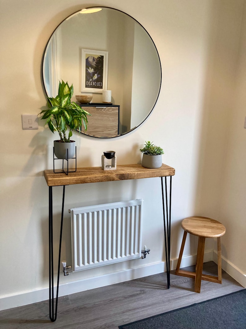 May include: A wooden console table with black hairpin legs, a round mirror with a black frame, and a white radiator. There are two potted plants and a small wooden stool on the table.