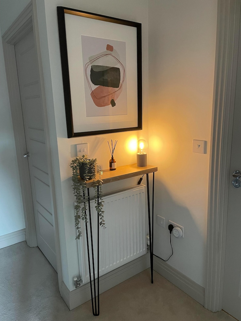K&ouml;nnte beinhalten: Ein schmaler Konsolentisch aus Holz mit schwarzen Haarnadelbeinen, einer Topfpflanze, einem Diffusor und einer Lampe mit einem klaren Glasschirm. Der Tisch steht in einer Ecke eines Raumes mit einem wei&szlig;en Heizk&ouml;rper und einer wei&szlig;en T&uuml;r.