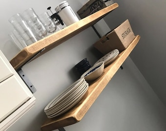 Puede incluir: Dos estantes de madera con soportes de metal negro montados en una pared blanca. El estante superior tiene una cafetera blanca, un frasco de vidrio y varias tazas de vidrio. El estante inferior tiene una pila de platos blancos, una cesta marrón etiquetada como "ALMACENAMIENTO" y varios cuencos azules y blancos.