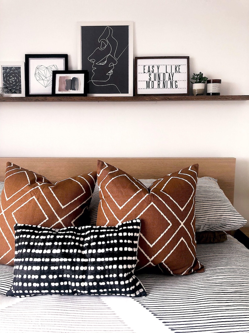 May include: A bed with a white and black striped duvet cover and brown and white patterned throw pillows. The pillows are arranged on a wooden headboard. Above the headboard is a shelf with black framed artwork and a sign that reads "EASY LIKE SUNDAY MORNING".