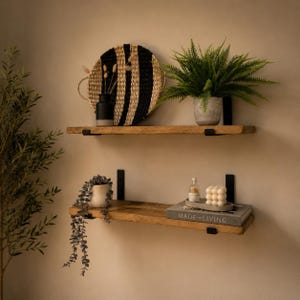 May include: Two natural wood floating shelves mounted with black metal brackets. The top shelf holds a black and tan woven circular tray, a black vase, and a green fern in a gray pot. The bottom shelf holds a trailing plant, a book titled MADE FOR LIVING, and a white bubble candle.