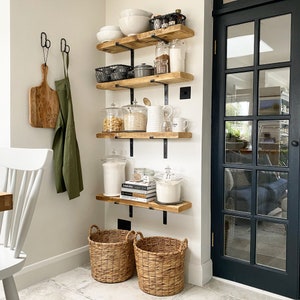 K&ouml;nnte beinhalten: Eine wei&szlig;e Wand mit vier Holzregalen mit schwarzen Metallkonsolen. Die Regale sind mit verschiedenen K&uuml;chenutensilien gef&uuml;llt, darunter Gl&auml;ser mit Pasta, Mehl und Zucker sowie Sch&uuml;sseln, Tassen und ein gusseiserner Topf. Unter den Regalen stehen zwei Weidenk&ouml;rbe auf dem Boden.