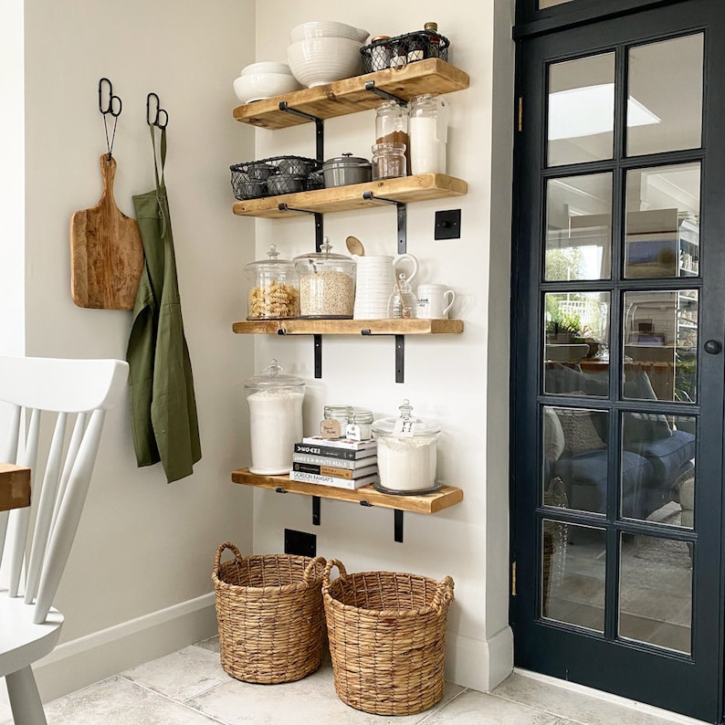 K&ouml;nnte beinhalten: Eine wei&szlig;e Wand mit vier Holzregalen mit schwarzen Metallkonsolen. Die Regale sind mit verschiedenen K&uuml;chenutensilien gef&uuml;llt, darunter Gl&auml;ser mit Pasta, Mehl und Zucker sowie Sch&uuml;sseln, Tassen und ein gusseiserner Topf. Unter den Regalen stehen zwei Weidenk&ouml;rbe auf dem Boden.