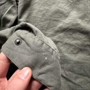 May include: Close-up of a section of a green garment, possibly a shirt or jacket. The fabric has a textured appearance with visible creases and folds. A button is visible on the cuff, and there are small white paint marks on the fabric.