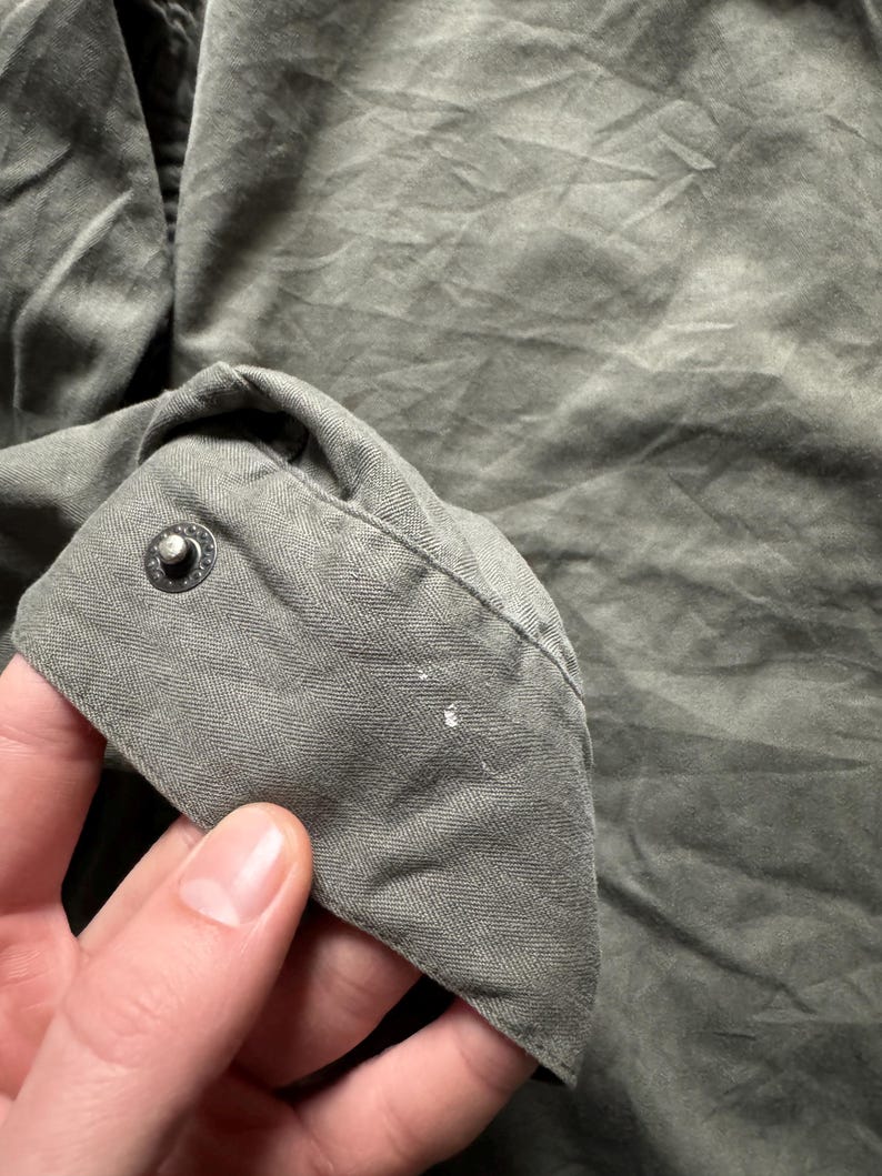 May include: Close-up of a section of a green garment, possibly a shirt or jacket. The fabric has a textured appearance with visible creases and folds. A button is visible on the cuff, and there are small white paint marks on the fabric.