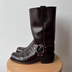 Puede incluir: Una bota de arnés de cuero marrón oscuro con punta cuadrada, caña a media pantorrilla y un detalle de anillo de metal plateado asegurado por correas de cuero y remaches en el empeine. La bota tiene un tacón bajo y costuras visibles.