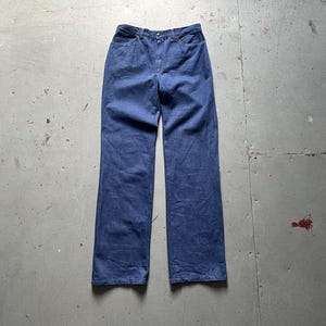 Puede incluir: Un par de pantalones vaqueros azules. Los pantalones tienen un diseño clásico de cinco bolsillos, cierre de botón y cremallera, y una pernera recta. El denim es de un lavado medio y los pantalones parecen estar en buen estado. Los pantalones están colocados sobre una superficie de hormigón.