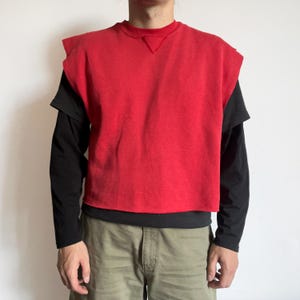 May include: A red sleeveless top with a crew neck, worn over a black long-sleeved shirt. The black sleeves extend beyond the red top. The person is wearing khaki shorts.
