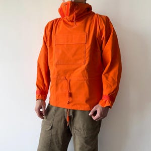 May include: Bright orange hooded anorak jacket with a large front pocket and drawstring adjustments. The jacket has long sleeves with red accents at the cuffs. The model is wearing olive green cargo pants.
