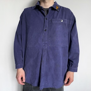 May include: A long-sleeved, navy blue shirt with a button-down collar and a single chest pocket. The shirt has a relaxed fit and features a small, yellow emblem on the collar. The shirt is made of a durable fabric.