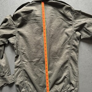 May include: Olive green jacket with a collared neckline and a buttoned closure. A measuring tape is placed along the center of the jacket, showing a length of approximately 30 inches. The jacket has a gathered hem and long sleeves.