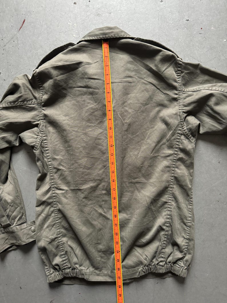 May include: Olive green jacket with a collared neckline and a buttoned closure. A measuring tape is placed along the center of the jacket, showing a length of approximately 30 inches. The jacket has a gathered hem and long sleeves.