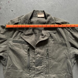 May include: Olive green military-style jacket with a collared neckline, two zippered pockets, and two velcro patches. The jacket is measured with a ruler, showing a length of approximately 17 inches.