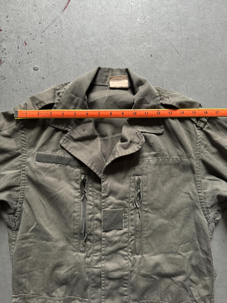 May include: Olive green military-style jacket with a collared neckline, two zippered pockets, and two velcro patches. The jacket is measured with a ruler, showing a length of approximately 17 inches.