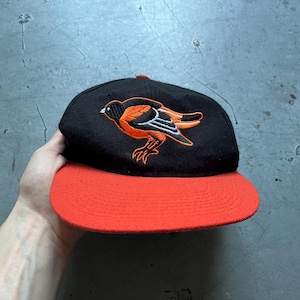 May include: Black baseball cap with a flat, orange brim. The cap features an embroidered orange and black bird logo on the front. A small red accent is visible on the top of the cap.
