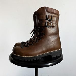 Puede incluir: Una bota de cuero marrón a media pantorrilla con cordones frontales, ojales metálicos y una correa ancha en el tobillo asegurada por dos hebillas de metal plateado. La bota tiene una suela de goma negra gruesa con costuras visibles.