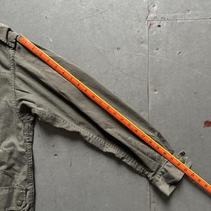 May include: Olive green jacket with a buttoned pocket and a ruler measuring the sleeve length. The ruler shows the sleeve is approximately 26 inches long. The jacket is made of a durable fabric.
