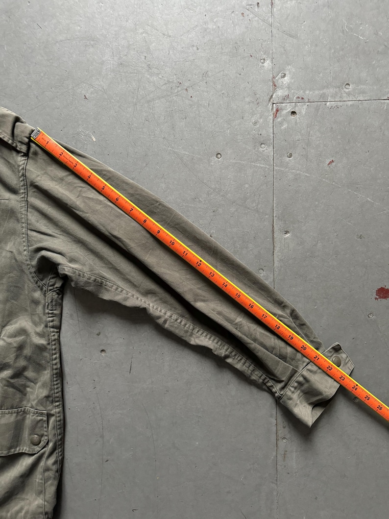 May include: Olive green jacket with a buttoned pocket and a ruler measuring the sleeve length. The ruler shows the sleeve is approximately 26 inches long. The jacket is made of a durable fabric.