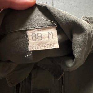 May include: Close-up of a military green garment tag. The tag displays the size "88 M" and the numbers "7080/8090". The fabric appears to be a durable, possibly cotton blend. The garment is olive drab in color.