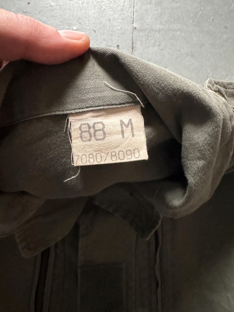 May include: Close-up of a military green garment tag. The tag displays the size "88 M" and the numbers "7080/8090". The fabric appears to be a durable, possibly cotton blend. The garment is olive drab in color.