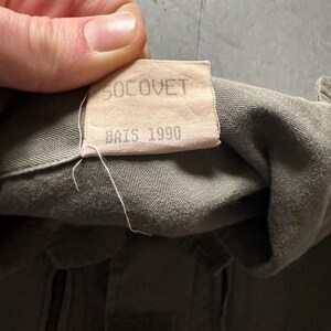 May include: Close-up of a tan label with the text "SOCOVET" and "BAIS 1990" printed in black. The label is attached to olive green fabric. The fabric appears to be part of a military garment.