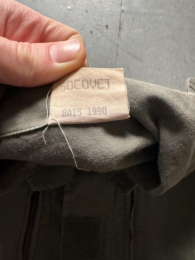 May include: Close-up of a tan label with the text "SOCOVET" and "BAIS 1990" printed in black. The label is attached to olive green fabric. The fabric appears to be part of a military garment.