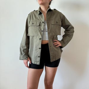 Può includere: Giacca verde oliva in stile militare con due tasche sul petto e chiusura con bottoni. La giacca ha maniche lunghe e spalline sulle spalle. La giacca è indossata aperta sopra un top grigio e pantaloncini neri.