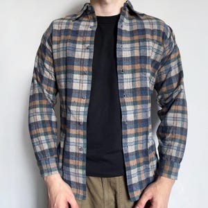 Puede incluir: Una camisa de cuadros con botones en tonos azules, beige y grises. La camisa de manga larga tiene un cuello clásico y se lleva sobre una camiseta negra. La camisa está hecha de una tela suave y tejida.