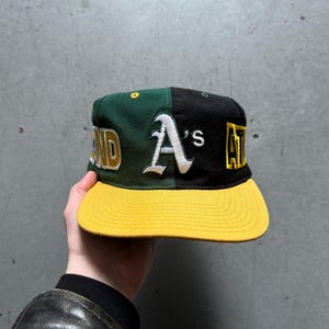 May include: A black and green baseball cap with a yellow brim. The front of the cap has the words "Oakland A's" embroidered in white.