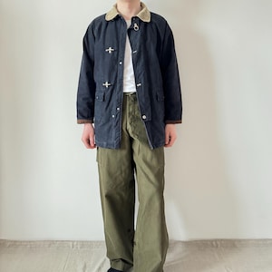 Puede incluir: Chaqueta azul marino con cuello de pana y cierres metálicos, combinada con pantalones cargo verde oliva. La chaqueta tiene un ribete marrón en los puños y el cuello. Un atuendo casual y práctico.