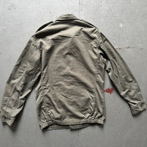 May include: Olive green jacket with a button-up front, long sleeves, and a collared neckline. The jacket has a gathered hem and cuffs, and is made of a textured fabric. The back of the jacket is visible.