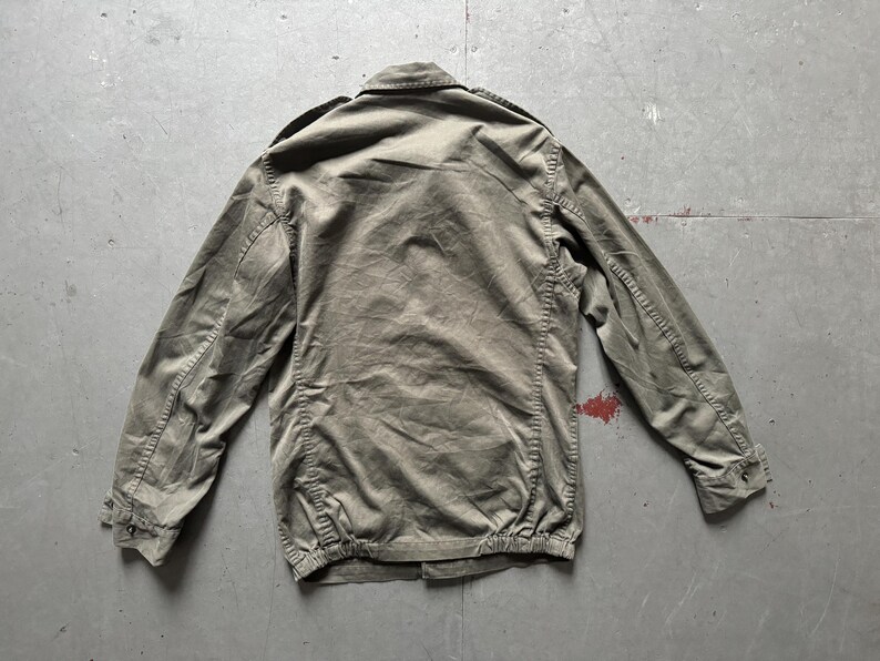 May include: Olive green jacket with a button-up front, long sleeves, and a collared neckline. The jacket has a gathered hem and cuffs, and is made of a textured fabric. The back of the jacket is visible.