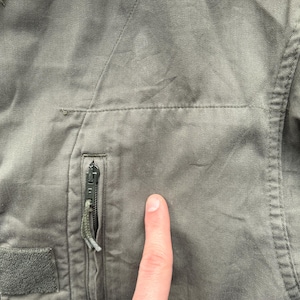 May include: Close-up of a faded olive green jacket. Visible features include a zippered pocket with a green pull, a velcro patch, and stitching details. The fabric appears worn and textured.