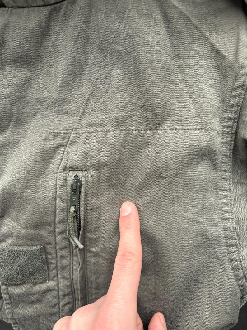 May include: Close-up of a faded olive green jacket. Visible features include a zippered pocket with a green pull, a velcro patch, and stitching details. The fabric appears worn and textured.