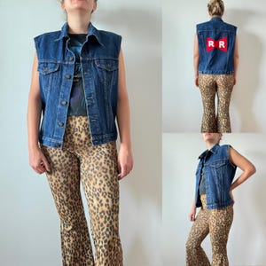 Può includere: Un gilet di jeans senza maniche con chiusura a bottoni e due tasche sul petto. La parte posteriore del gilet presenta una grafica rossa e bianca. Il gilet è abbinato a pantaloni con stampa leopardata.