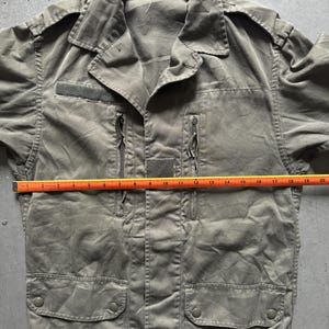 May include: Olive green military-style jacket with a collared neckline, two zippered pockets, and two flap pockets. The jacket has a small patch on the chest and a tag at the collar. The jacket measures approximately 20 inches across the chest.