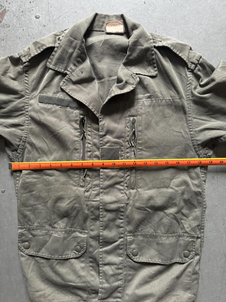 May include: Olive green military-style jacket with a collared neckline, two zippered pockets, and two flap pockets. The jacket has a small patch on the chest and a tag at the collar. The jacket measures approximately 20 inches across the chest.
